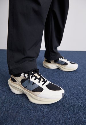 Hvide og sorte sneakers med strukturerede overflader og blå detaljer, der har en kraftig sål og sorte snørebånd, parret med sorte bukser.