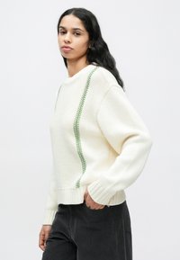 Bijeli pleteni džemper s teksturiranim završetkom, sadrži zelenu prugu duž bočnih šavova i rebraste manžete i rub. Uparen s tamnim hlačama.