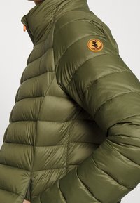 Groene pufferjack met gewatteerde textuur, ritssluiting en oranje accent op de kraag. Bevat een ronde patchlogo op de bovenarm.