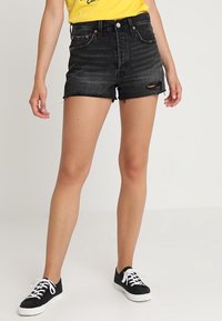 Shorts de mezclilla negros de talle alto con dobladillos deshilachados, detalles desgastados y herrajes de tono cobre, combinados con zapatillas negras sobre un fondo liso.