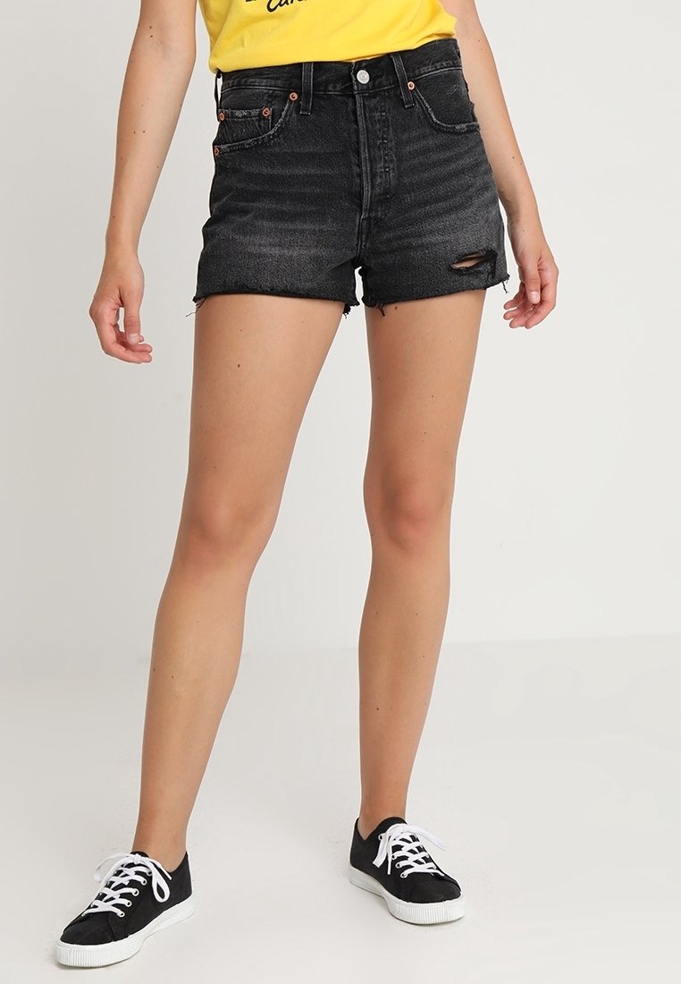 Shorts de mezclilla negros de talle alto con dobladillos deshilachados, detalles desgastados y herrajes de tono cobre, combinados con zapatillas negras sobre un fondo liso.