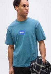 Άνδρας που φοράει μπλουζάκι HUGO σε τιρκουάζ χρώμα, κοιτάζει δεξιά, κρατώντας μαύρο-άσπρο καρό μπουφάν με το αριστερό χέρι σε ουδέτερο φόντο.