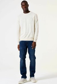 Man staat in een crèmekleurige gebreide trui, donkerblauwe jeans en witte sneakers tegen een lichte effen achtergrond.