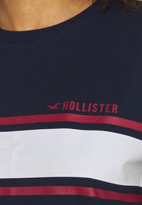 Granatowa koszulka z okrągłym dekoltem w poziome czerwone i białe paski, z logo "HOLLISTER" w kolorze czerwonym umieszczonym nad paskami.