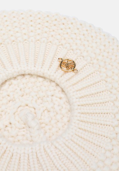 Béret tricoté couleur crème avec motifs en côtes et point popcorn, et un petit emblème décoratif métallique rond doré.