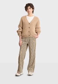 Cardigan a maglia beige con cinque bottoni, abbinato a pantaloni con stampa leopardata e sneakers di colore chiaro. Il modello è in piedi davanti a uno sfondo neutro.