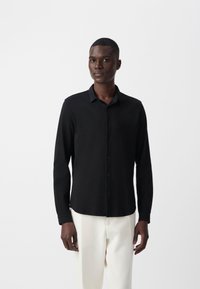 Camisa negra de manga larga hecha de tela suave, con botonadura delantera, cuello estructurado y dobladillo recto, combinada con pantalones claros.