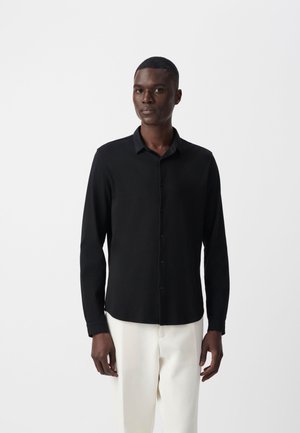 Hombre de pie con una camisa negra de manga larga y pantalones blancos frente a un fondo blanco liso.