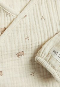 Beige musliini kangas, jossa on pieniä ruskeita karhu- ja lehtikuvioita, teksturoidulla pinnalla. Sisältää taitetun hihansuun, jossa on etiketti.