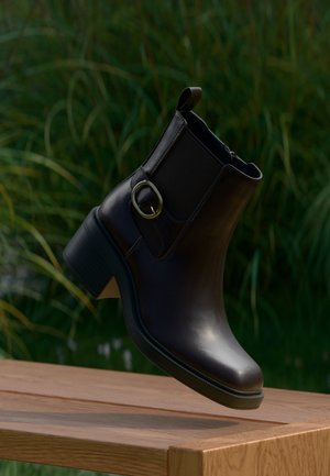 Schwarzer Leder-Stiefelette mit niedrigem Absatz und seitlicher Schnalle, die über einer Holzoberfläche im Freien mit grünem Laub im Hintergrund schwebt.