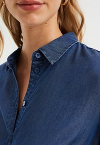 Koszula jeansowa o miękkiej fakturze, ciemnoniebieskim kolorze, kołnierzyku na guziki i dwoma niebieskimi guzikami przy szyi, w klasycznym kroju.