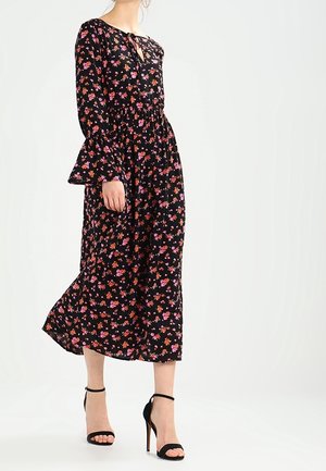 Femme portant une robe midi noire à motifs floraux roses et orange avec manches longues, associée à des sandales noires à talons hauts.