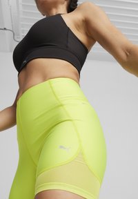 Juoda sportinė liemenėlė ir ryškiai neoniniai geltoni šortai su tinklu. Šortai turi subtilų logotipą ir sklandžią, elastingą medžiagą.