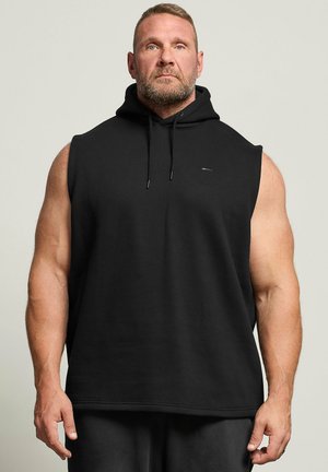 Spierbundige man met baard staat frontaal, draagt een zwarte mouwloze hoodie en zwarte broek tegen een egale lichte achtergrond.