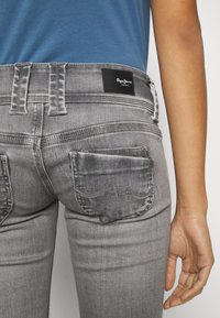 Jeans de mezclilla gris con un diseño ajustado, que presentan dos bolsillos traseros y una etiqueta de marca negra en la cinturilla. Textura suave visible.