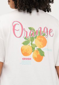 Inimene, kellel on seljas valge t-särk, millel on värvikas oranž graafika, kus peal on sõna "Oranž" silmatorkavalt välja toodud.