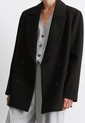 Persoon draagt een zwarte dubbelgeknoopte jas over een grijs gestreept overhemd met knopen en bijpassende broek.