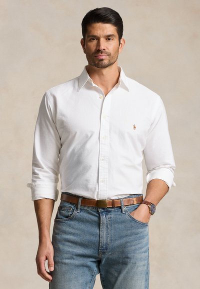 Mand med kort mørkt hår iført hvid skjorte med knapper, blå jeans, brunt bælte og armbåndsur, stående med den ene hånd i lommen.