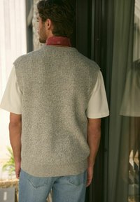 Szary dzianinowy kamizelka z teksturowanym wzorem, krótki rękaw w jasnym beżu, noszona na koszuli z kołnierzykiem. Luźny krój, prosty design.