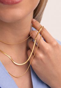 Tre catene metalliche d'oro di diverse lunghezze sono sovrapposte attorno a un collo. Le catene hanno una texture liscia e una superficie riflettente.