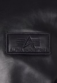 Melna ādas birka ar iepresātu logotipu "ALPHA INDUSTRIES" ar stilizētām līnijām un leņķotu dizainu. Redzamas teksturētas virsmas detaļas.