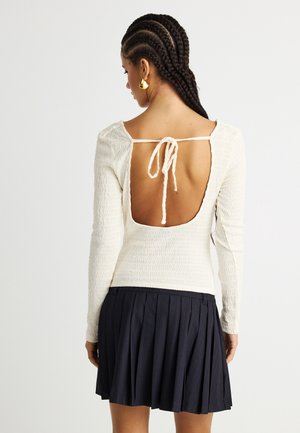 Γυναίκα με πλεξούδες, που φορά χρυσά σκουλαρίκια, λευκό μακρυμάνικο μπλουζάκι με υφή και ανοιχτή πλάτη δεμένη με κορδόνια, και μαύρη φουστα με πιέτες.