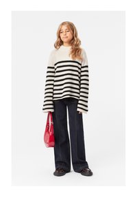 Prugasti pulover u krem i crnoj boji, široki rukavi, uparen sa tamnim raširenim trapericama. Model drži sjajnu crvenu torbicu i nosi crne cipele.