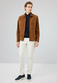 Bruine suede jas met een kraag, voorrits en zijzakken, gecombineerd met een zwarte polo, crèmekleurige broek en marineblauwe sneakers.