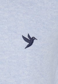 Jasnoniebieska dzianina z ciemnoniebieskim haftowanym logo kolibra, umiejscowionym centralnie po lewej stronie. Miękka tekstura, prosty design.