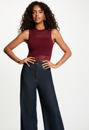 Vrouw met krullend haar, die een mouwloos bordeaux topje en een hoge taille brede, donkere spijkerbroek draagt, staand met handen in de zij tegen een witte achtergrond.