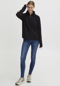 Pullover nero a costine con colletto con zip, polsini larghi e vestibilità rilassata, abbinato a jeans skinny blu e sneakers sportive di colore chiaro.