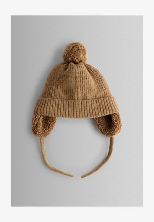 Brūna adīta cepure ar ribotu dizainu, pūkainu pom-pomu virsotnē un ausu atloci, kas izklāta ar mīkstu, teksturētu audumu. Iespējo pievilkšanas saitītes drošai piegulšanai.