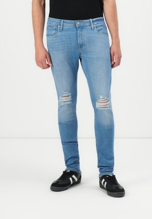 Light blue skinny jeans with distressed knee rips; classic five-pocket design; paired with black suede sneakers featuring white accents.