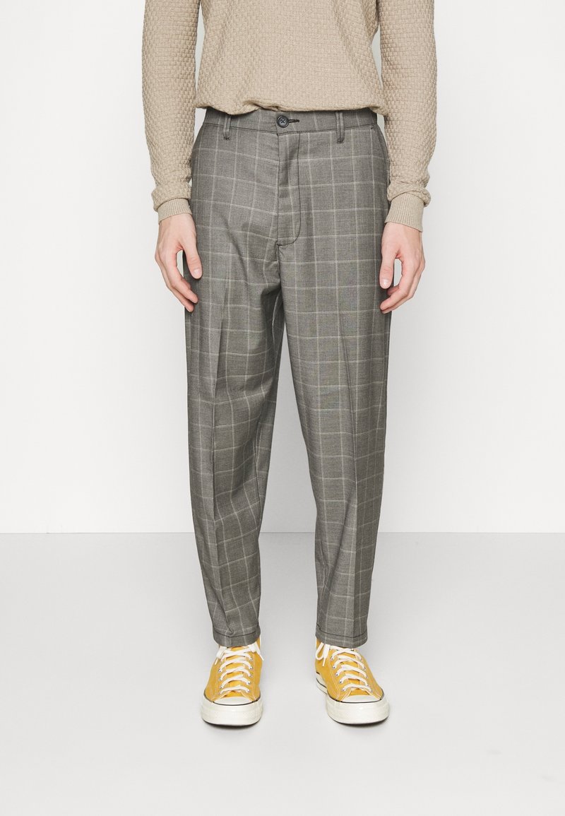 Homme portant un pantalon gris à carreaux, un pull beige texturé et des baskets jaunes, debout devant un fond blanc.