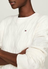 Biały sweter w splot warkoczowy z prążkowanym okrągłym dekoltem, z małym, haftowanym logo na piersi. Sweter jest lekki i ma miękką teksturę.