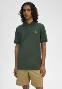 Polo verde con colletto e maniche corte, dotato di un logo ricamato dorato sul petto, abbinato a pantaloni cargo color khaki.