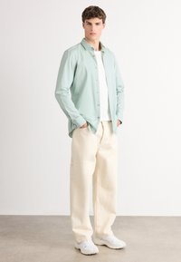 Cămașă butonată de culoare verde deschis cu guler, tricou alb dedesubt, pantaloni largi de culoare crem și adidași albaștri. Design minimalist, textură netedă.