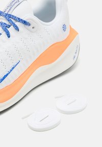 Weißer Sportschuh mit blauen Schnürsenkeln, orangefarbener Zwischensohle, strukturiertem Obermaterial. Zwei zusätzliche weiße Schnürsenkel in der Nähe positioniert.