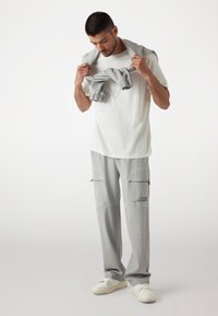 Gray pants with side zippered pockets and logo detail, paired with a white t-shirt. The model holds a gray sweater draped around the neck.