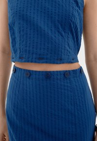 Modrý texturovaný dvoudílný outfit, který zahrnuje krátký top s kulatým výstřihem a sukní s vysokým pasem, zdobenou pěti knoflíky.