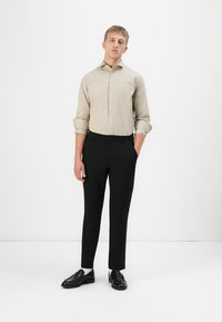 Jonge man staat in een beige knoopshirt met opgerolde mouwen, zwarte op maat gemaakte broek en zwarte instappers tegen een witte achtergrond.