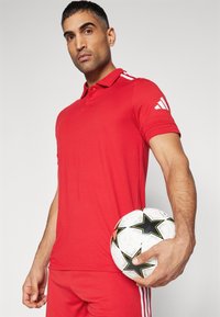 Rode polo met een kraag en drie knopen, met witte strepen op de mouwen. De persoon houdt een witte voetbal met zwarte sterren vast.