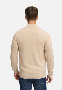 Beige stickad tröja med hög krage, ribbstickade ärmslut och nederkant, samt en mjuk textur. Bärs över mörkblå denimjeans.