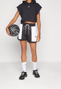 Hoodie cropped senza maniche nera con logo, abbinata a pantaloni sportivi nero e bianco. Sneakers nere e calze bianche completano l'outfit.