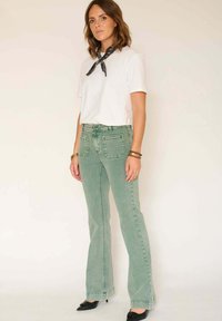 Jean évasé vert avec une finition en denim doux et texturé, doté de deux poches avant et de poches plaquées à l'arrière. Associé à un T-shirt blanc.