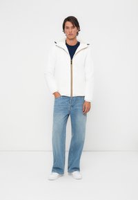 Hvid zip-jakke med gule detaljer, der har hætte og blød foring, parret med lyseblå jeans og hvide sneakers.