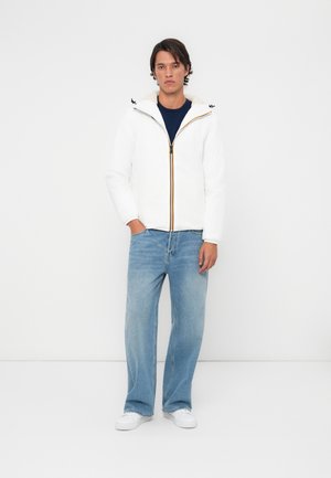 Witte zip-up jas met gele accenten, voorzien van een capuchon en zachte voering, gecombineerd met lichtblauwe jeans en witte sneakers.