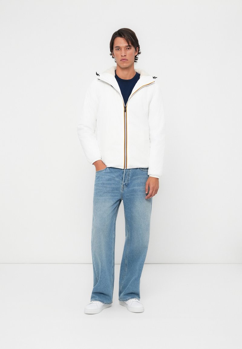 Hvid zip-jakke med gule detaljer, der har hætte og blød foring, parret med lyseblå jeans og hvide sneakers.