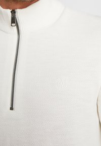 Maglione bianco con zip e colletto alto, realizzato in tessuto a maglia testurizzato e con un logo discreto in rilievo sul petto. Dettaglio della zip argentata.