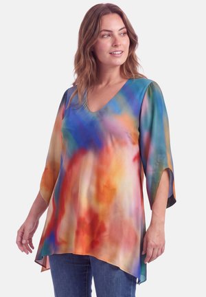 Vrouw die een kleurrijke tie-dye tuniek draagt met driekwartssplitmouwen en een V-hals, gecombineerd met een blauwe spijkerbroek, staand tegen een effen achtergrond.